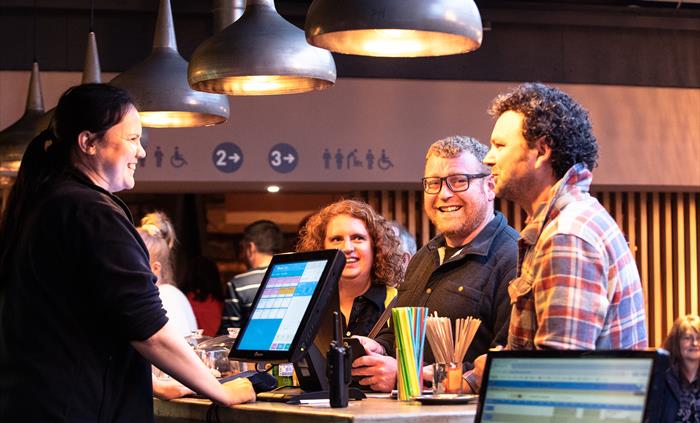 A man with a checked shirt, a man with glasses and a woman with curly hair stand at a bar being served by a lady with a dark ponytail